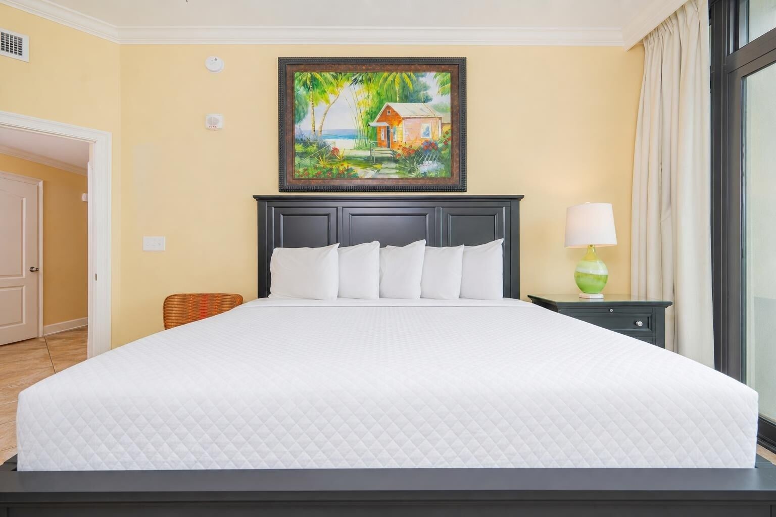 A hotel room with a large bed, a painting above the bed, a nightstand with a lamp, and a view of the outdoors through a window.