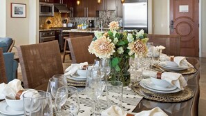 A dining area with a table set for six, a vase of flowers, and a kitchen in the background. - Honua Kai | Ocean View 3 BR Sleeps 8 | Car Incl. w/ 6+ Nights | HKH-925 by KBM (Lahaina)