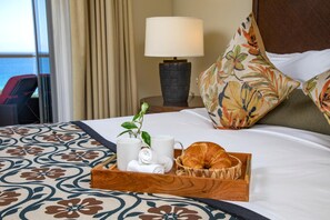 Ein ordentlich bezogenes Bett mit einer blumigen Tagesdecke, ein Tablett mit einem Croissant, einer Tasse und einer Pflanze.