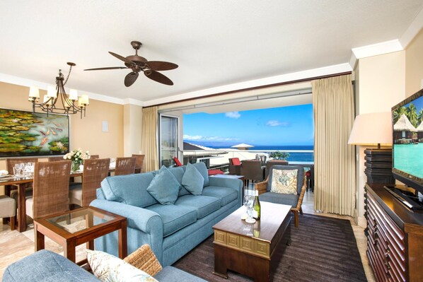 Ein Wohnzimmer mit blauem Sofa, Esstisch und Stühlen, einem Deckenventilator und einem großen Fenster mit Meerblick.