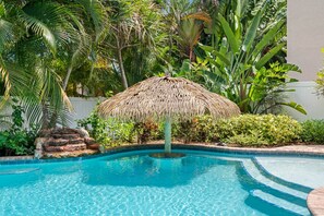 A swimming pool with a thatched umbrella, surrounded by tropical plants and a stone structure.