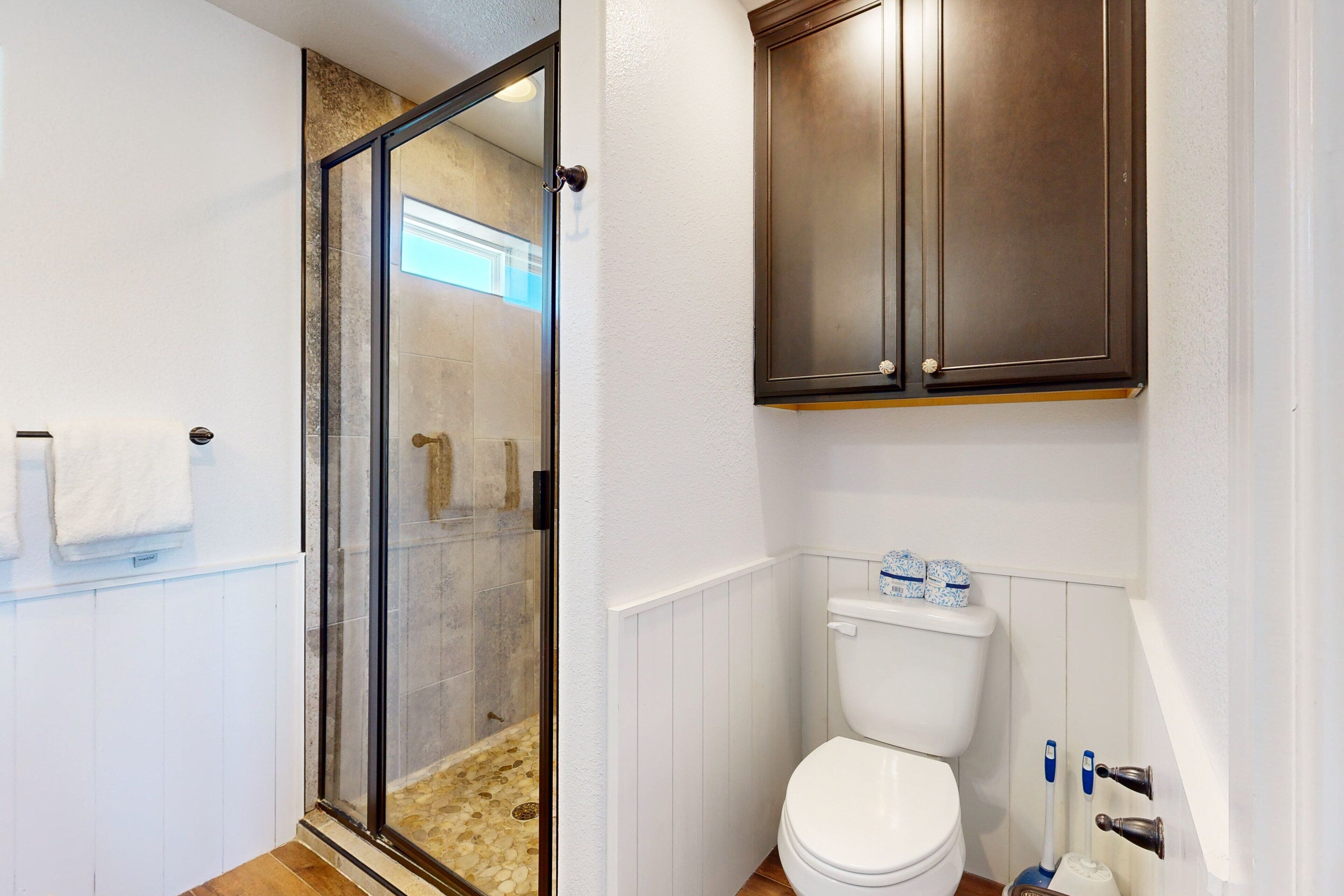 Un baño con cabina de ducha de vidrio, inodoro blanco y armarios de madera sobre el inodoro.