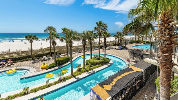 Un área de piscina con varias piscinas, palmeras y una playa al fondo.