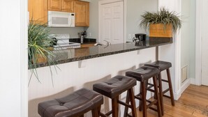 A kitchen with wooden cabinets, a white refrigerator, a microwave, and a sink. There are three stools and a potted plant on the counter. - Palmetto Perch - Fully Stocked Wild Dunes Condo with Pool, Close to Beach (Isle of Palms)