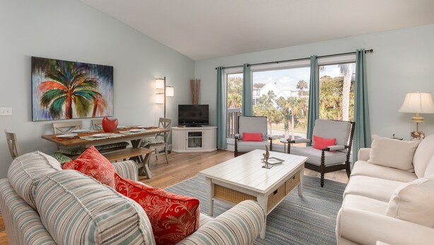 A living room with a dining area, a TV, and large windows with curtains. - Palmetto Perch - Fully Stocked Wild Dunes Condo with Pool, Close to Beach (Isle of Palms)
