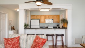 A living room with a striped sofa, two red patterned pillows, a ceiling fan, and a kitchen area with wooden cabinets and a refrigerator. - Palmetto Perch - Fully Stocked Wild Dunes Condo with Pool, Close to Beach (Isle of Palms)