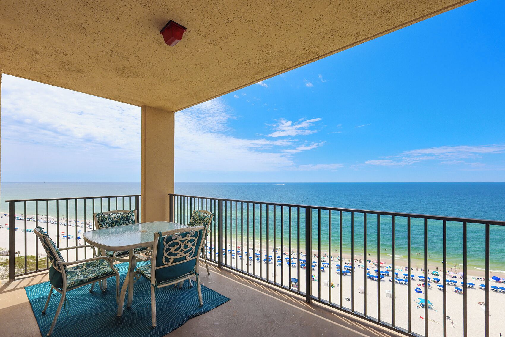 A balcony with a table and chairs, overlooking a beach with umbrellas and people.
