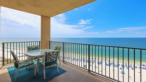 Un balcon avec une table et des chaises, donnant sur une plage parsemée de parasols et fréquentée par des gens.