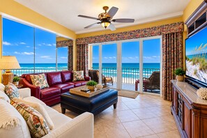 A spacious living room with a red leather sofa, a blue ottoman, and a television on a wooden cabinet. Large windows offer a view of the ocean. - Direct Oceanfront * Heated Pool & Hot Tub * 3BR + Recharge Lounge (Daytona Beach Shores)