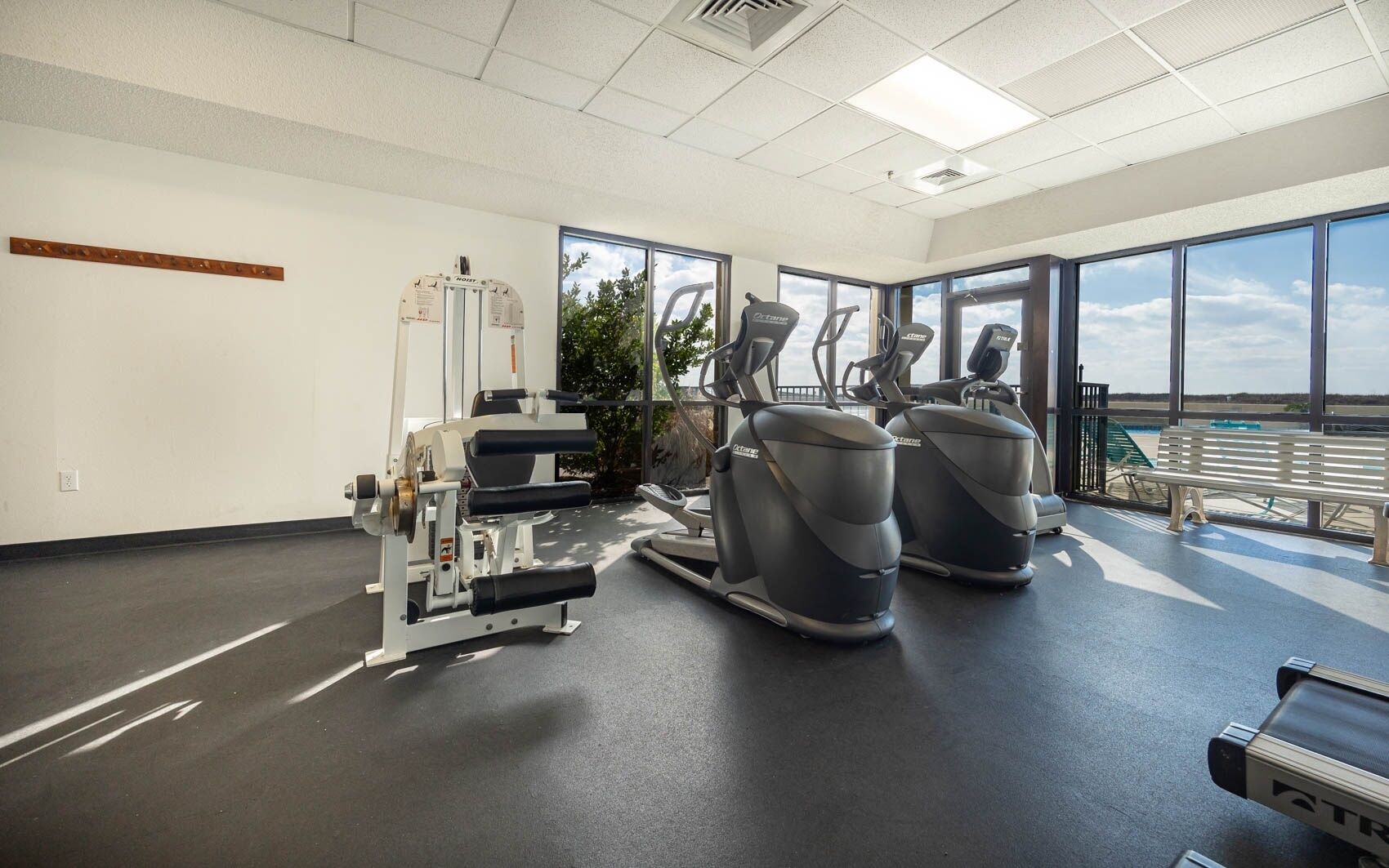 A gym with exercise equipment, large windows, and a bench.