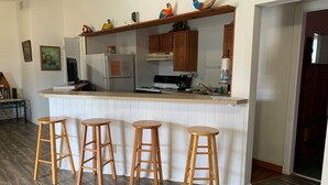 A kitchen with wooden stools, a refrigerator, cabinets, and a shelf with decorative items.