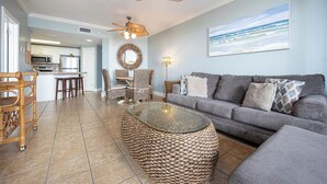 A living room with a grey sofa, wicker coffee table, and wicker chairs. There is a kitchen in the background with white cabinets and a microwave.
