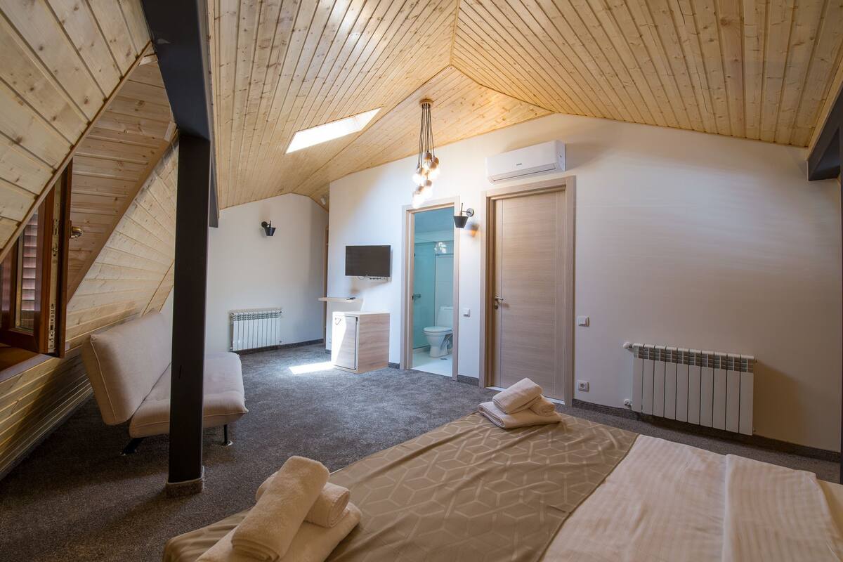 A bedroom with a sloped ceiling, a bed, a small bedside table, a wall-mounted TV, and a bathroom visible through an open door.