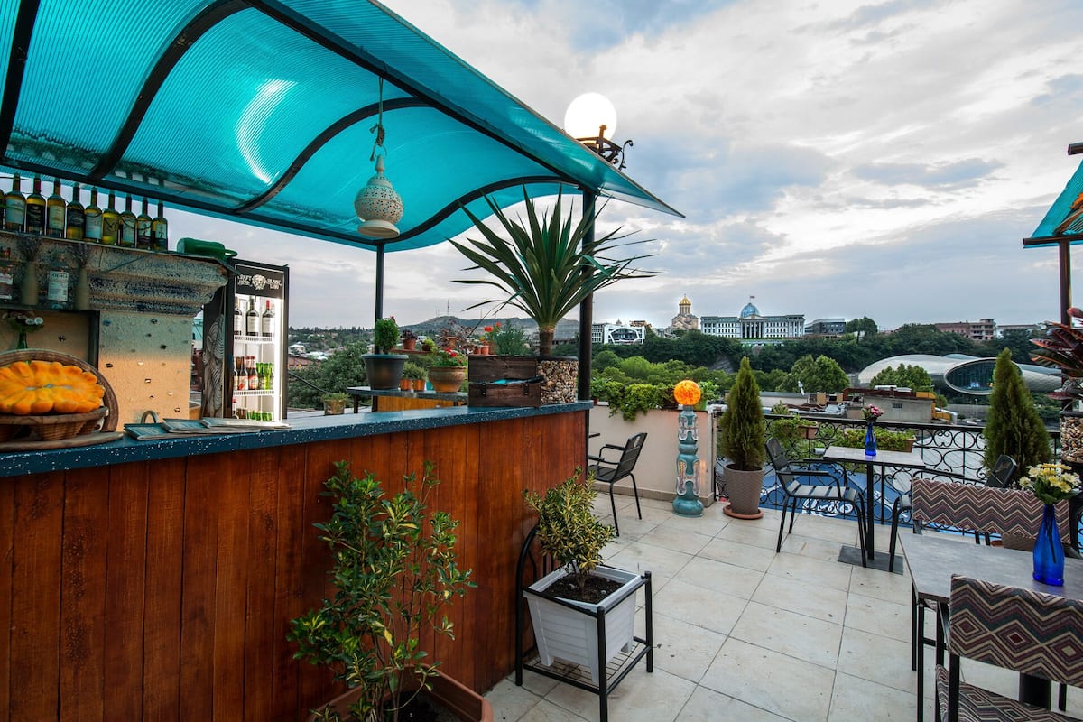 A rooftop bar with a wooden counter, potted plants, and a view of city buildings.