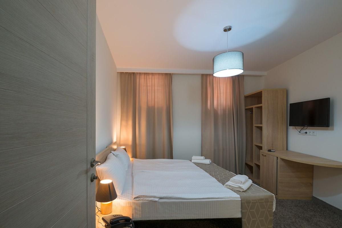 A hotel room with a bed, bedside lamp, curtains, a TV, and a wooden wardrobe.
