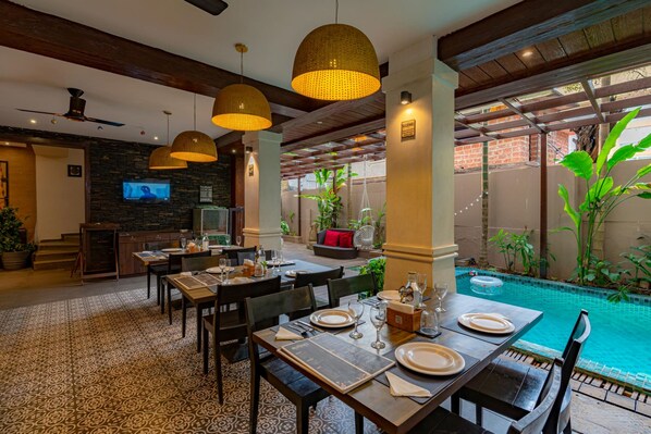 A restaurant with outdoor seating, a pool, and a TV screen. - Riva Gold Coast (Candolim)