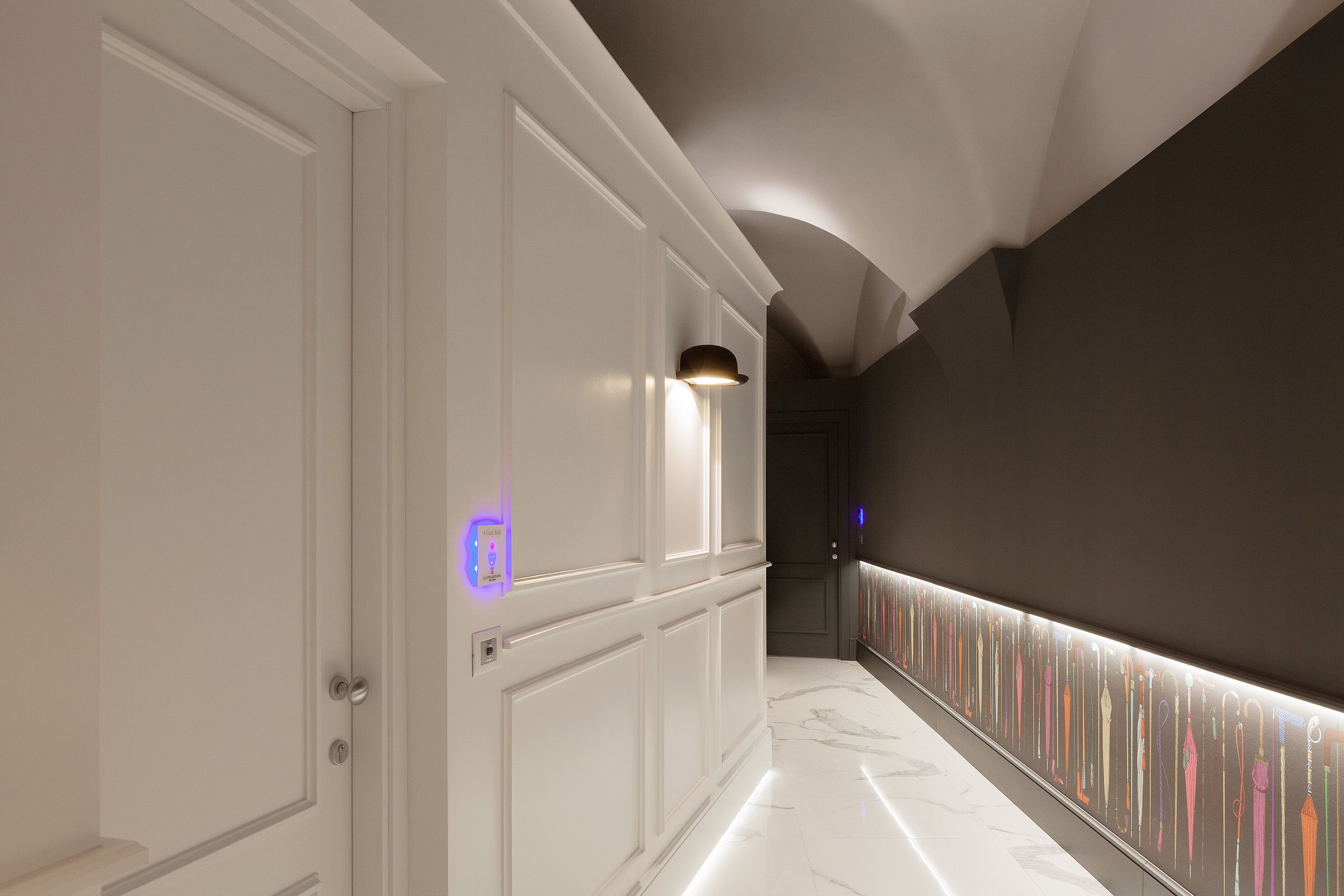 Un couloir aux murs recouverts de panneaux blancs, doté d’un sol en marbre et d’un mur orné de divers objets.