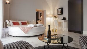 A modern bedroom with a curved bed, a glass coffee table with a bottle and glasses, and a bedside lamp. - Quattro Canti Suites (Catania)