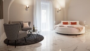 A modern bedroom with a round bed, a houndstooth armchair, a small round table with a bottle and glasses, and a large window with sheer curtains. - Quattro Canti Suites (Catania)