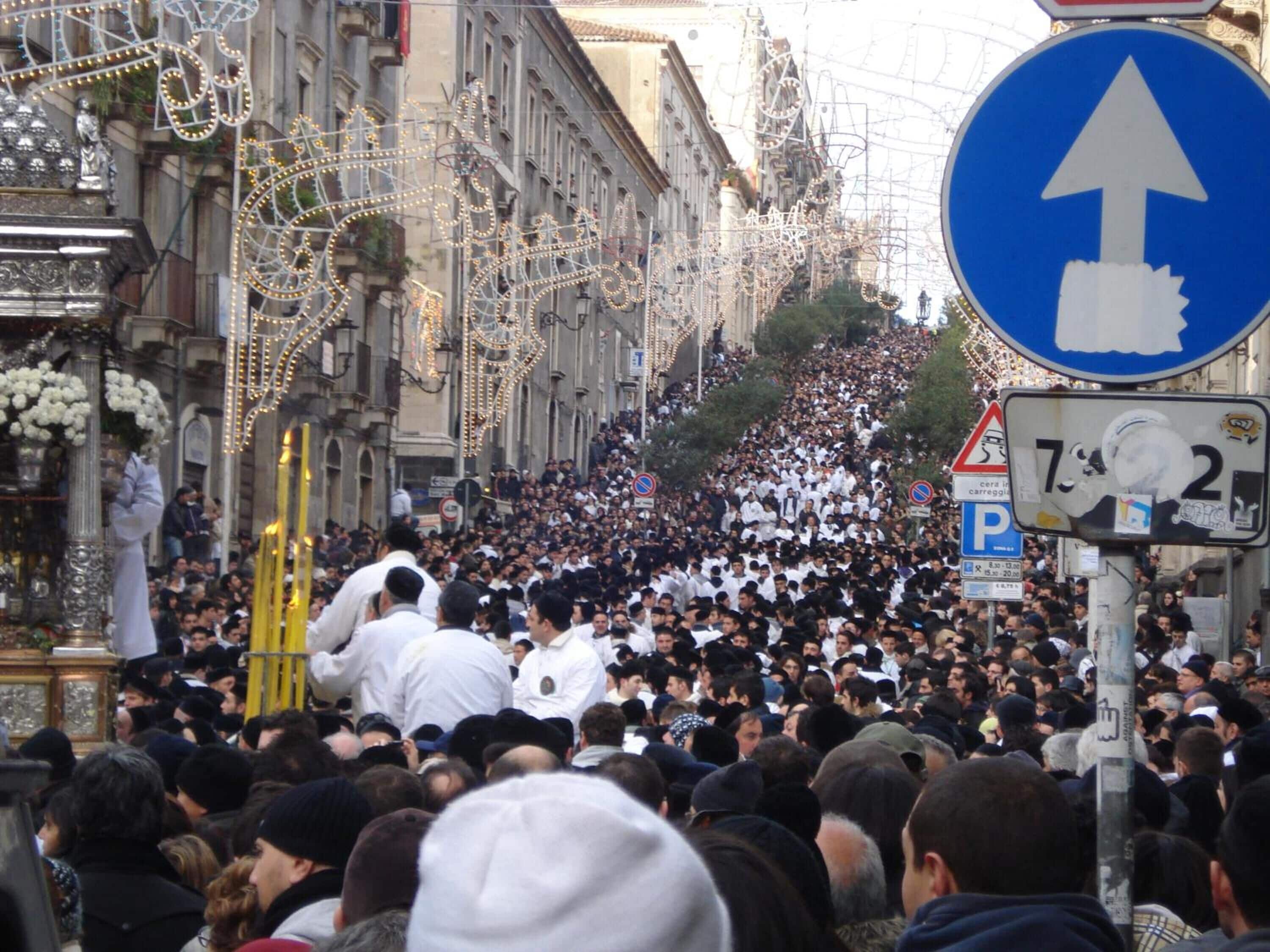 Calle concurrida con un gran grupo de personas, algunas llevando objetos religiosos, y una señal azul direccional.