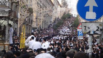 Calle concurrida con un gran grupo de personas, algunas llevando objetos religiosos, y una señal azul direccional.