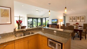 A modern kitchen with granite countertops, wooden cabinets, and a built-in oven. There is a dining area with a table and chairs, and a large window offering a view of the outdoors. - Spacious Modern Haven Just Steps from the Beach - Wailea Elua #908 -Lush Grounds (Wailea, Maui)