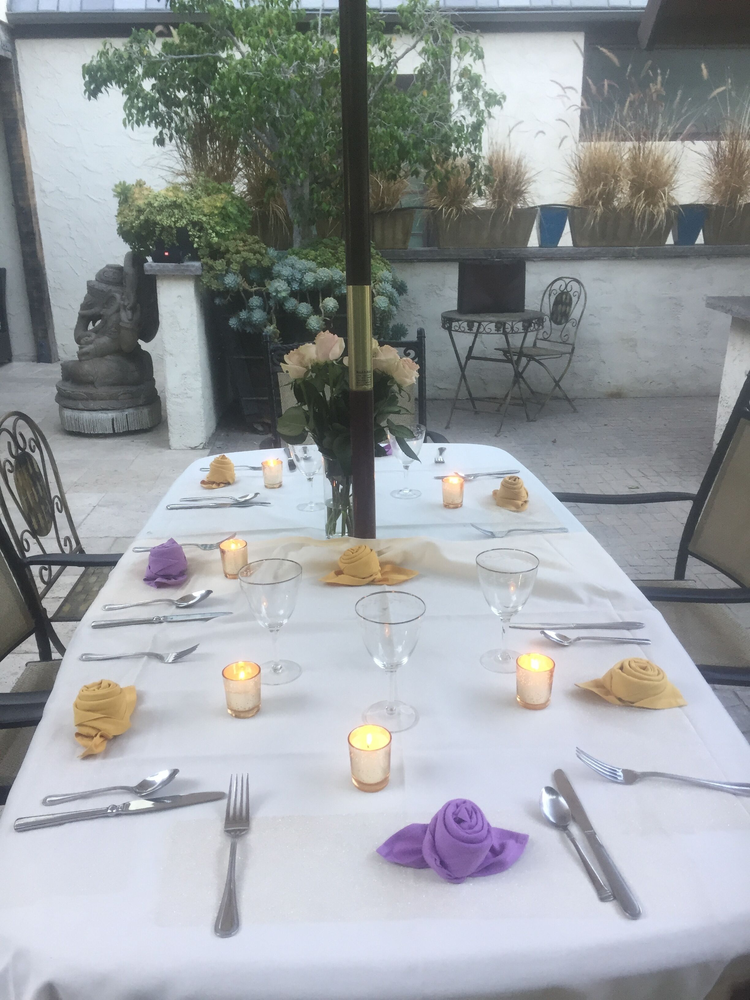 Una mesa preparada para una comida, con velas, una estatua y plantas en macetas al fondo.