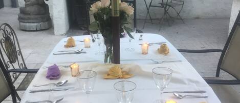 Una mesa preparada para una comida, con velas, una estatua y plantas en macetas al fondo.