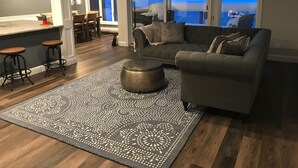 Uma sala de estar moderna com sofá cinza, tapete estampado e piso de madeira.