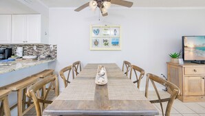 A dining area with a wooden table, chairs, a ceiling fan, and a kitchen in the background.