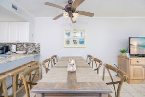 A dining area with a wooden table, chairs, a ceiling fan, and a kitchen in the background.