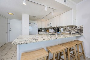 A kitchen with white cabinets, a granite countertop, a stainless steel sink, and wooden bar stools.