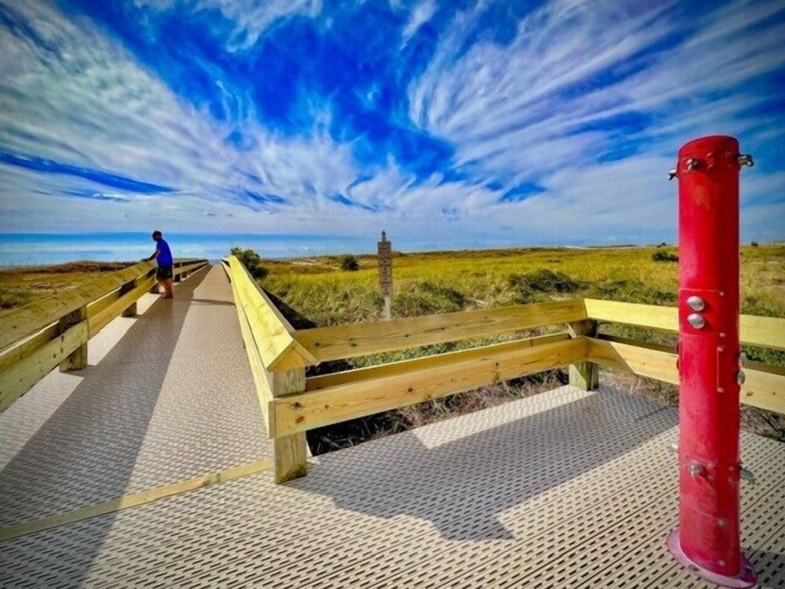A wooden boardwalk leading to a beach with a red post on the right and a person in blue at the start.