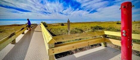 A wooden boardwalk leading to a beach with a red post on the right and a person in blue at the start.