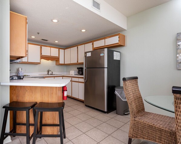 A compact kitchen with wooden cabinets, a stainless steel refrigerator, a small island with stools, and a wicker chair.