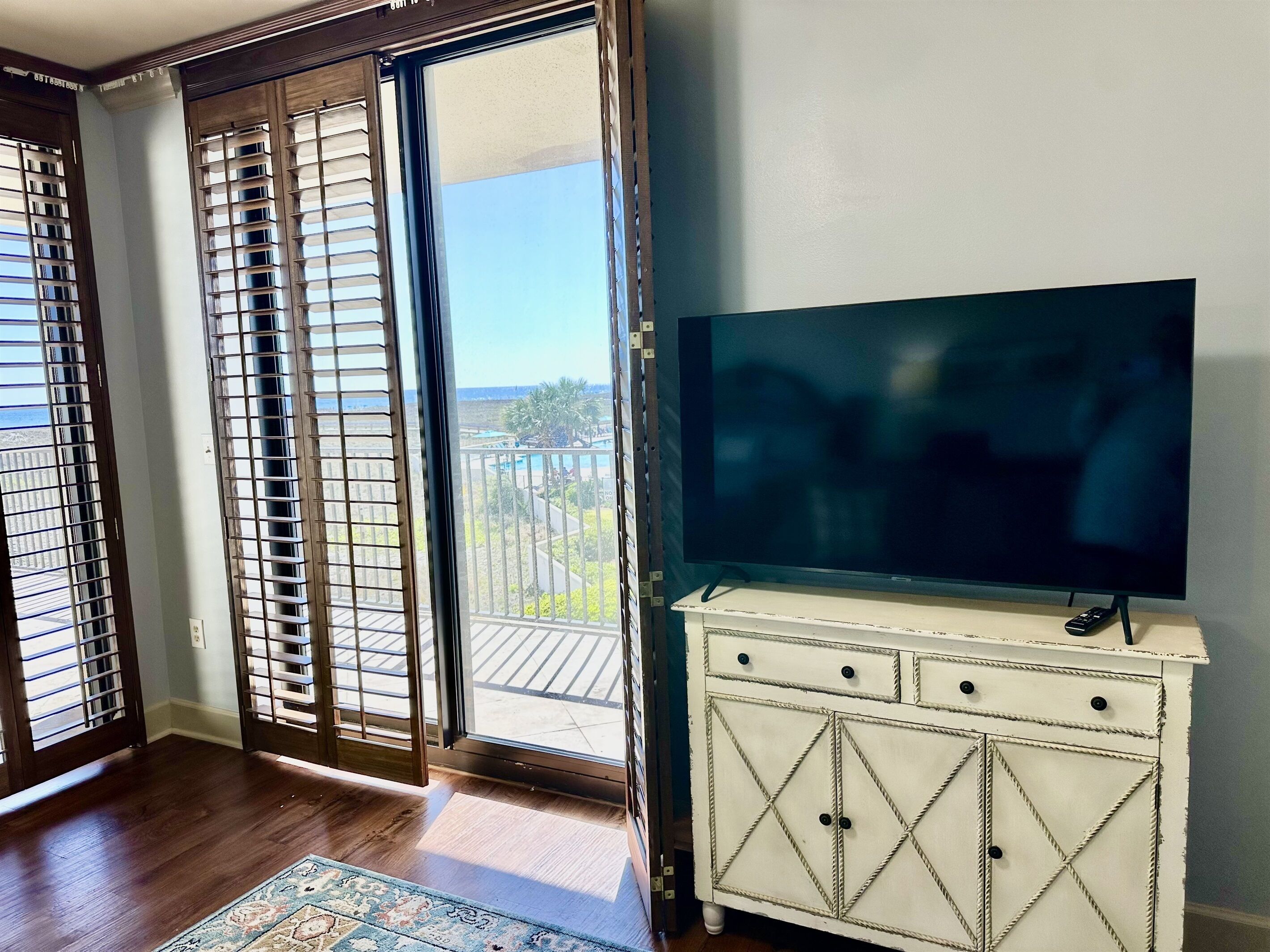 A room with a sliding glass door leading to a balcony, a flat-screen TV on a white cabinet, and a patterned rug on the floor.