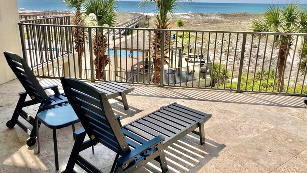 Balcón con dos sillas negras y una pequeña mesa, con vista a una playa y una zona de piscina.