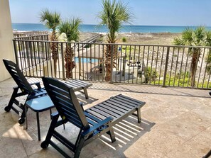 A balcony with two black chairs and a small table, overlooking a beach and a pool area.
