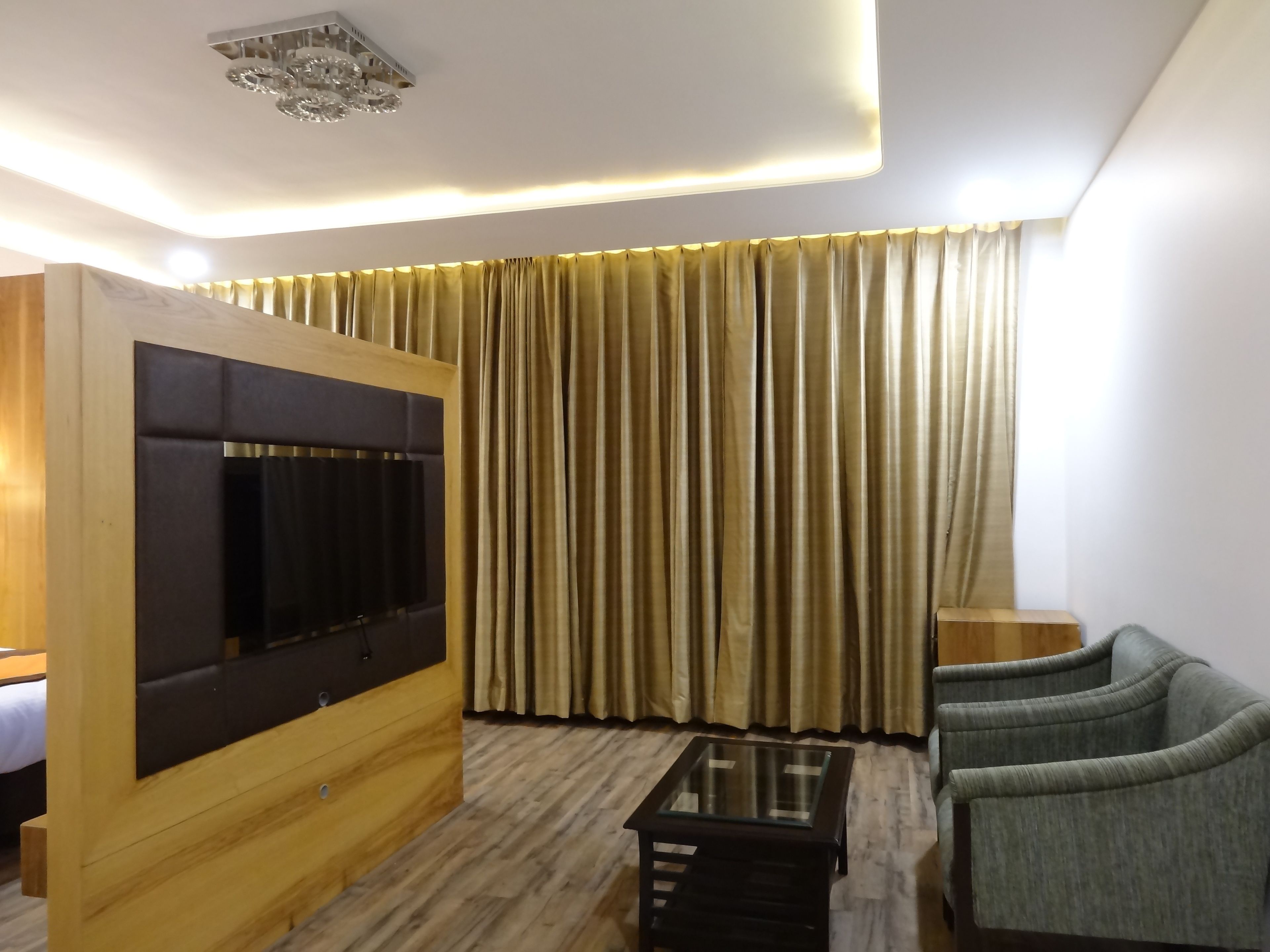 Moderni hotellihuone, jossa on puinen sängynpääty, lasinen sohvapöytä, kaksi verhoiltua tuolia ja suuri ikkuna, jossa on kullanväriset verhot.