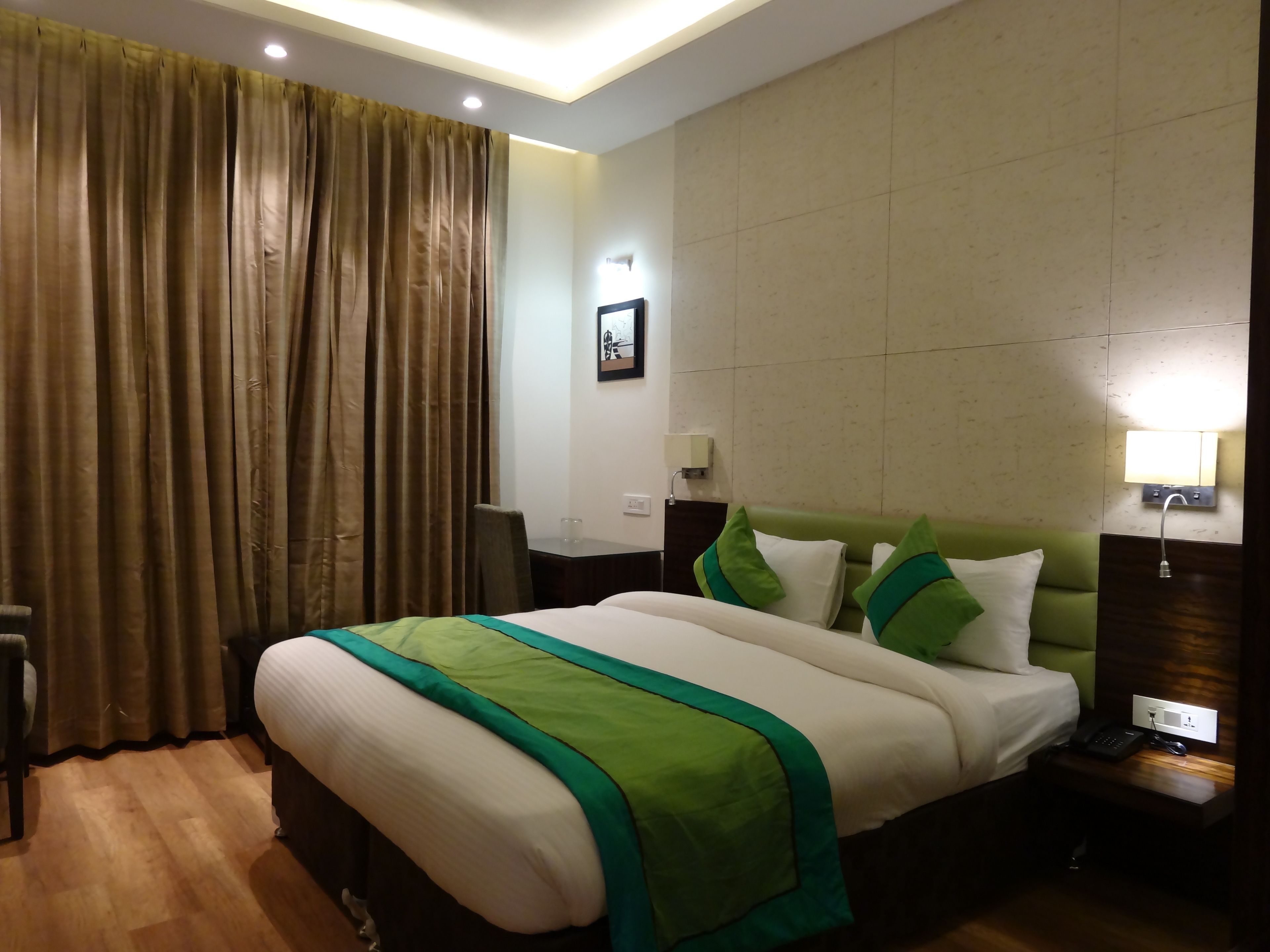 A hotel room with a large bed, a green headboard, a nightstand with a lamp, and a window with curtains.