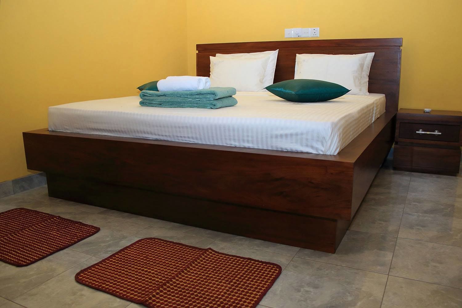 A large bed with a wooden headboard, white bedding, and a green pillow. There are two brown mats on the floor.