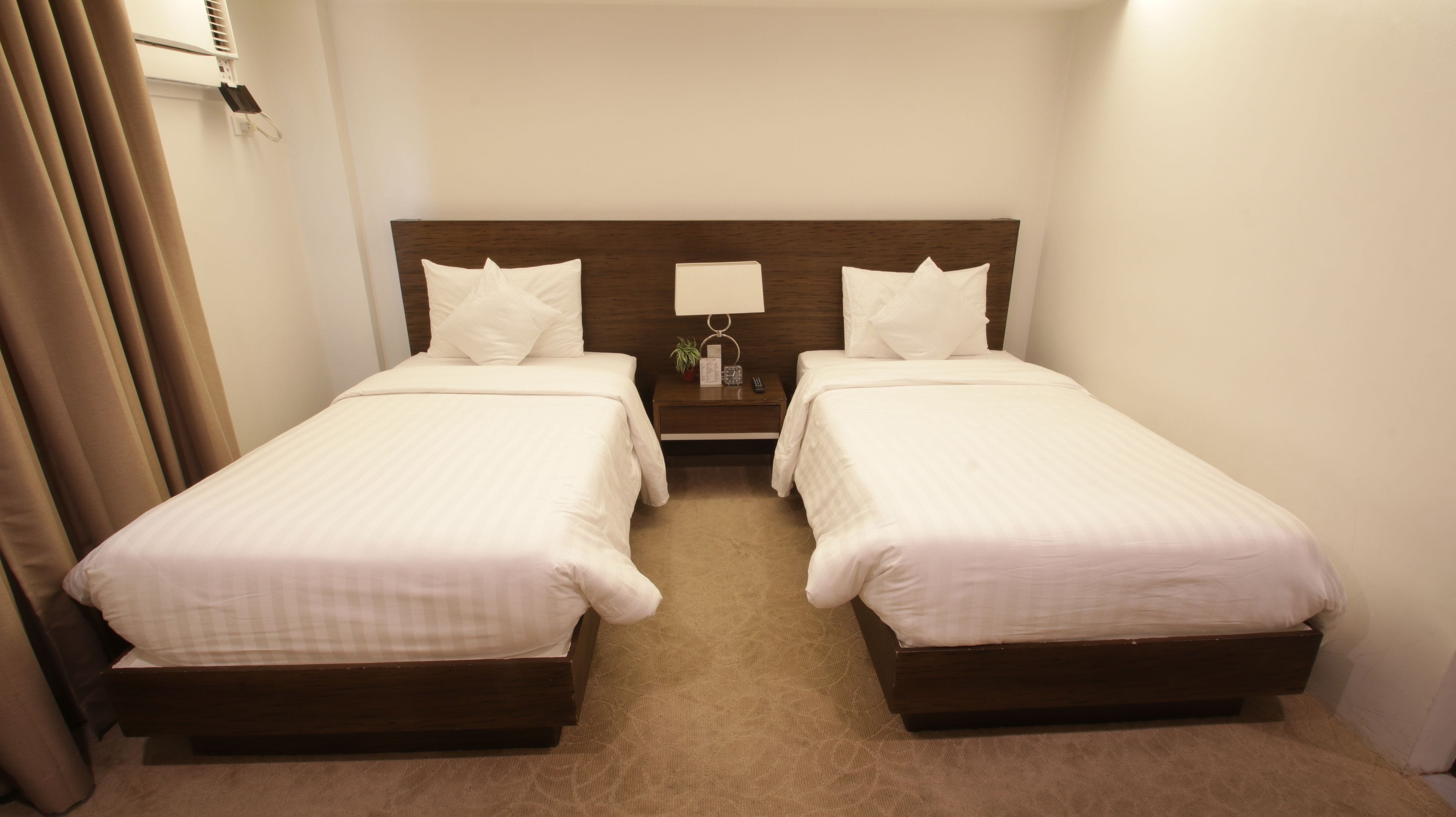 A hotel room with two single beds, a nightstand with a lamp, a remote control, and a small plant.