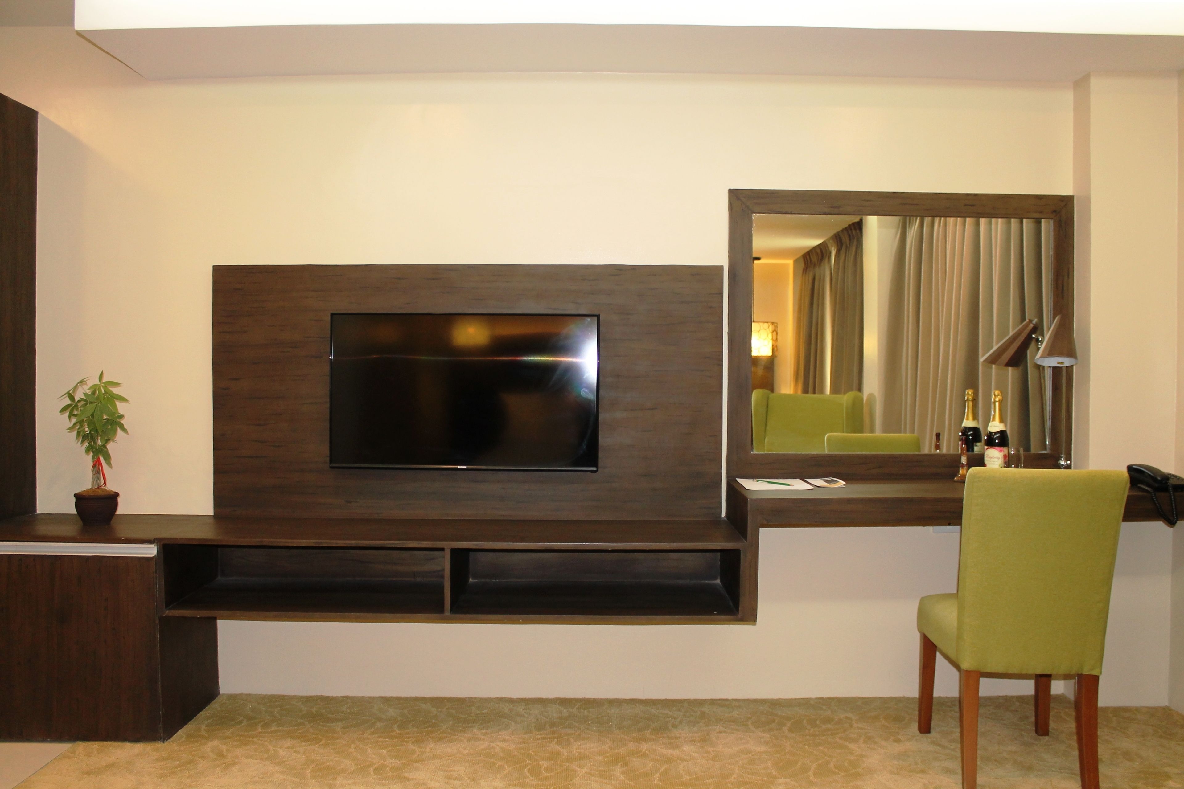 A modern hotel room with a flat-screen TV mounted on a dark wood panel, a green chair, a small potted plant, and a mirror reflecting the room.