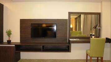 A modern hotel room with a flat-screen TV mounted on a dark wood panel, a green chair, a small potted plant, and a mirror reflecting the room.