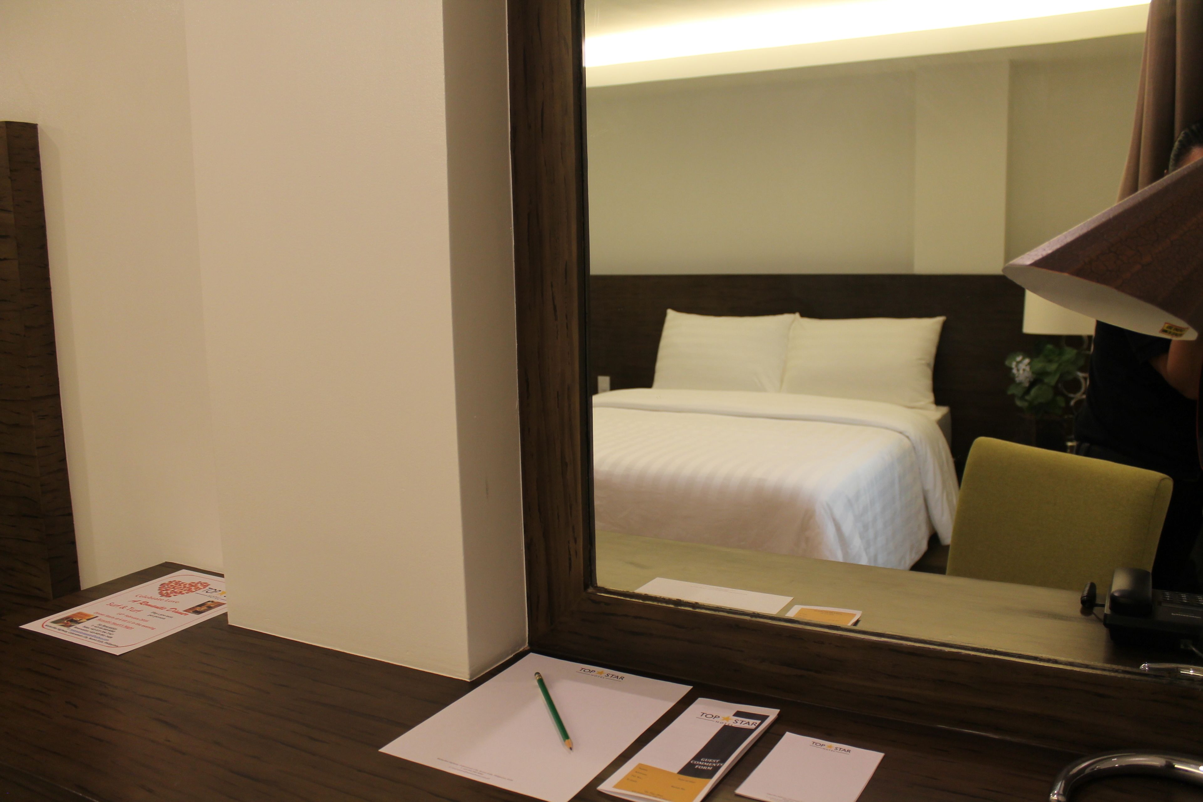 A hotel room with a large mirror reflecting a bed and a desk with a lamp, papers, and a pen.