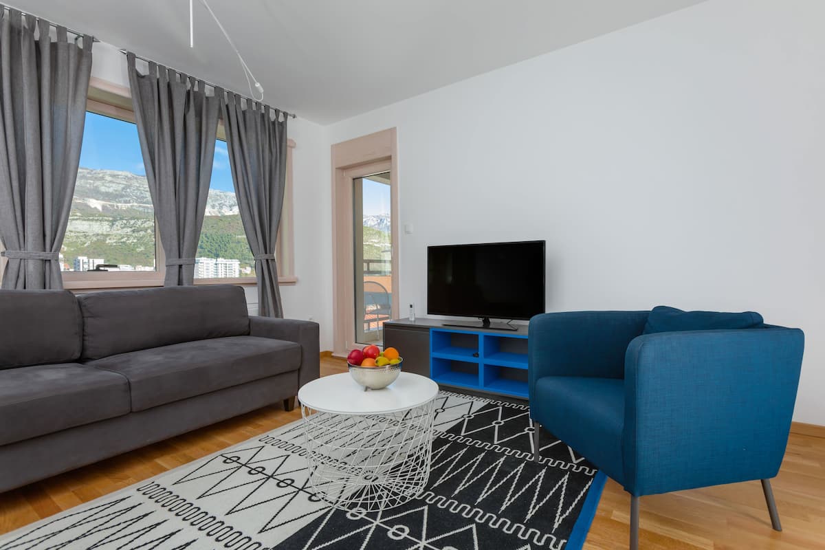 A modern living room with a grey sofa, a blue armchair, a flat-screen TV, and a bowl of fruit on a coffee table.