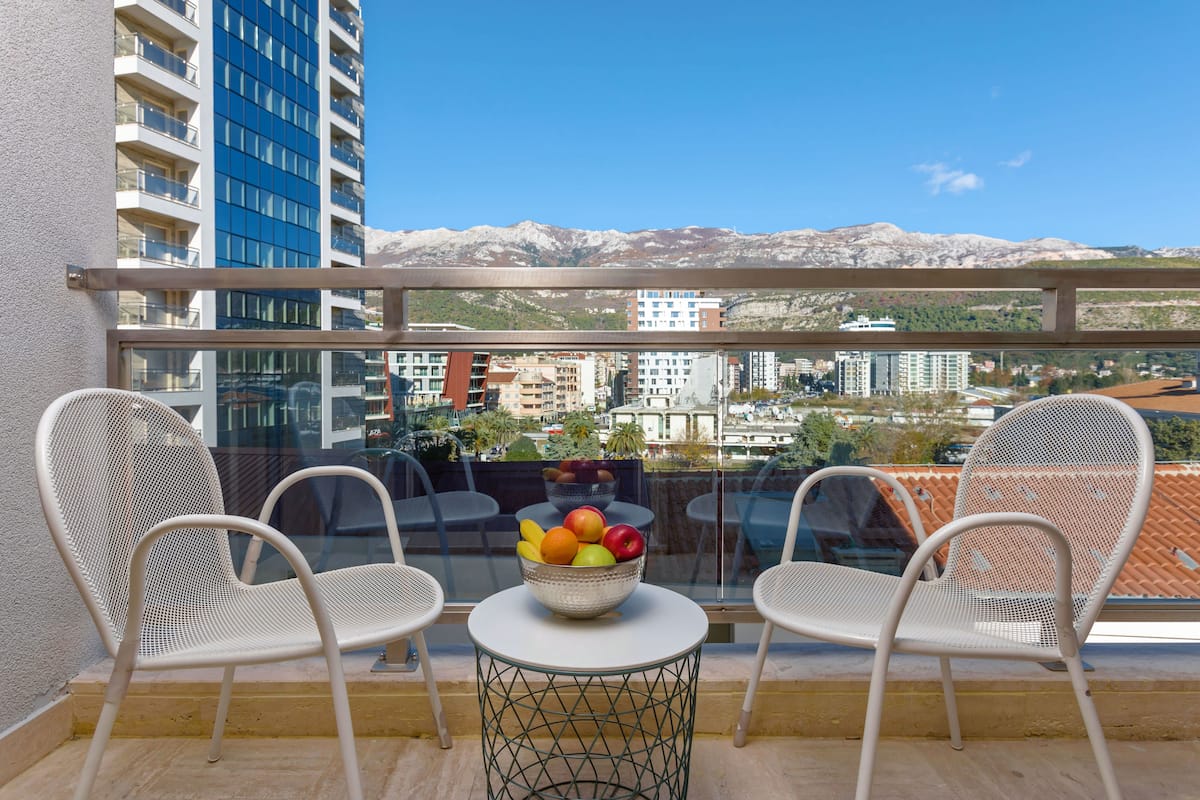 A balcony with two white mesh chairs, a small round table, and a bowl of fruit. A cityscape with buildings and mountains is visible in the background.