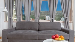 A grey sofa in a room with a large window, a white table with a fruit bowl, and a lamp. - Apartments Krug (Budva)