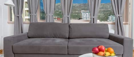 Um sofá cinzento numa divisão com uma janela grande, uma mesa branca com uma fruteira e um candeeiro.