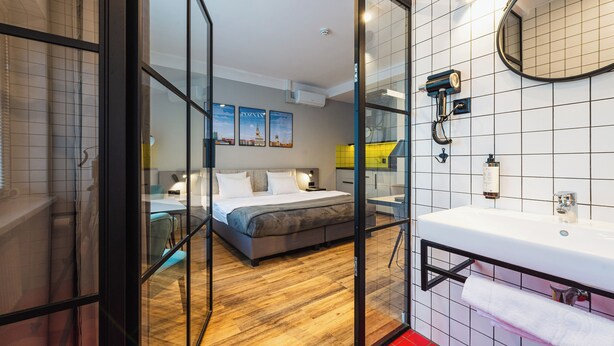 Et moderne hotelværelse med trægulv, en stor seng, en glasvæg, et badeværelse med hvid vask og spejl, samt bybilleder på væggen.